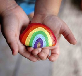The hands of a young toddler child are gently holding an acrylic painted rainbow rock.